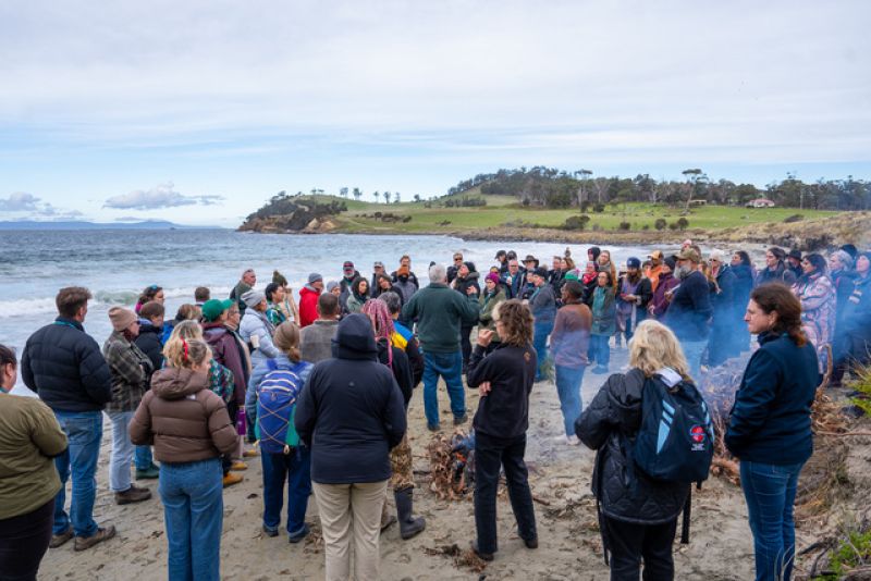 Large group of people standing around a fire on the beach listening to a speaker in the centre.
