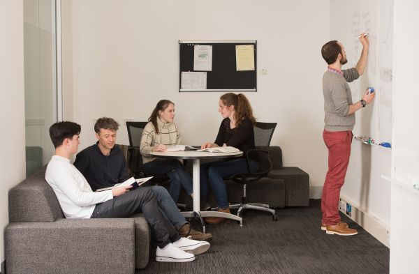 Five people in a quiet study space - some are sitting on a couch, some are chatting at a table, and one is writing on a white board