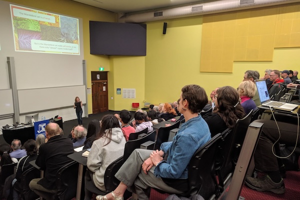 A lecture hall full of people of all ages, listening to a woman at looking at a screen displaying Mandelbrot sets